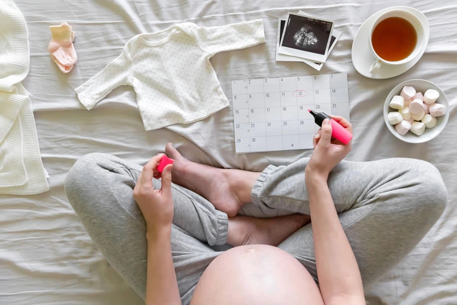 Trudna devojka sedi u turskom sedu na krevetu i zapisuje u kalendar trudnoce.