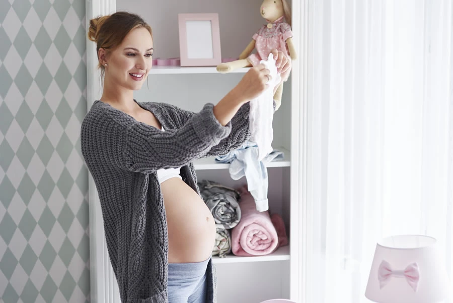 Trudna devojka stoji u decijoj sobi i drzi garderobu za bebe.