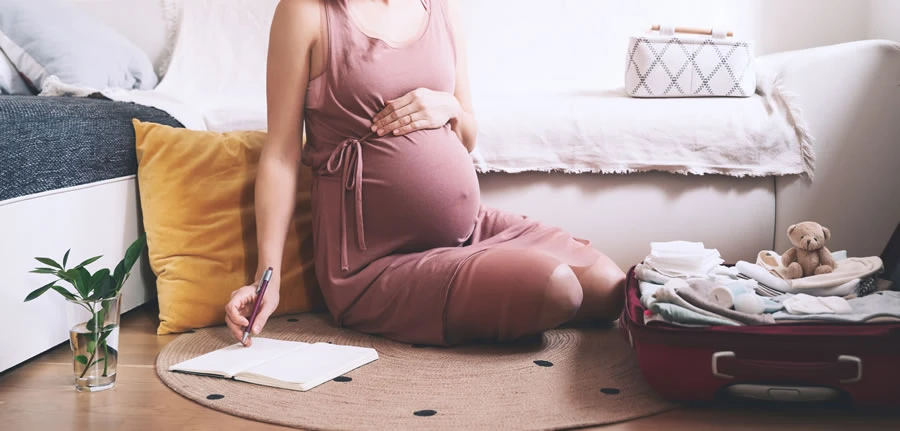 Trudna devojka u 32. nedelji trudnoce sedi na podu pokraj kreveta i zapisuje u svesku.